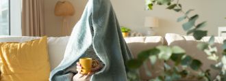 Eine Person sitzt mit einem Tee auf dem Sofa und hat sich eine Decke über den Kopf gezogen.