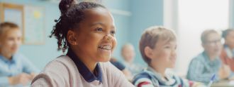 Ein Mädchen sitzt in der Schule an ihrem Platz und lächelt, wobei ihre Zahnspange gut zu erkennen ist. 