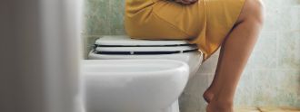 Eine Frau sitzt angezogen auf einer Toilette und hält sich den Bauch vor Schmerzen.