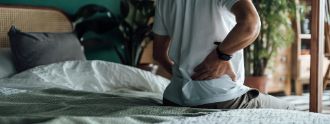 Ein Mann sitzt auf dem Bett und hält sich die Hände an seinen unteren Rücken.
