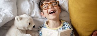 Lachender Junge mit Brille liegt im Bett und liest ein Buch.