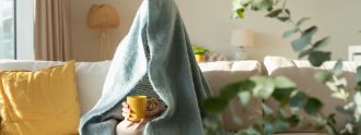 Eine Person sitzt mit einem Tee auf dem Sofa und hat sich eine Decke über den Kopf gezogen.
