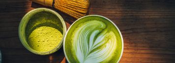 Man sieht einen Becher mit Matcha, sowie einen Matcha Besen und weiteres Zubehör.