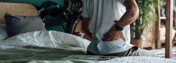 Ein Mann sitzt auf dem Bett und hält sich die Hände an seinen unteren Rücken.