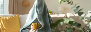 Eine Person sitzt mit einem Tee auf dem Sofa und hat sich eine Decke über den Kopf gezogen.