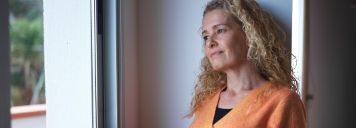 Eine mittelalte Frau steht mit einem Tee am Fenster und schaut raus.