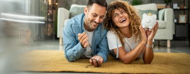 Ein paar liegt auf dem Boden mit Geld und einem Sparschwein in der Hand und freut sich.