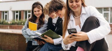 Vier Jugendliche sitzen nebeneinander, das Mädchen im Vordergrund schaut auf ihr Smartphone und laechelt.