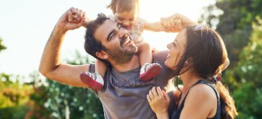 Vater trägt Kleinkind auf Schultern, Mutter schaut beide an, alle lachen und sind fröhlich.