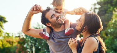 Vater trägt Kleinkind auf Schultern, Mutter schaut beide an, alle lachen und sind fröhlich.