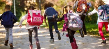 Grundschulkinder mit Schulranzen rennen draußen auf einem Weg und sind von hinten zu sehen.