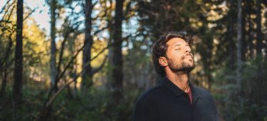 Ein Mann steht entspannt im Wald und genießt den Sonnenstrahl und die innere Ruhe.