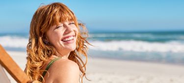 Frau sitzt auf Klappstuhl aus Holz am Strand und lacht glücklich in die Kamera. 