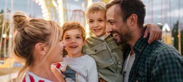 Mutter und Vater stehen sich glücklich auf einem Jahrmarkt gegenüber, jedes Elternteil trägt einen Jungen im Kindesalter auf dem Arm.