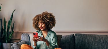 Eine glückliche Frau, die auf ihrem Handy schreibt, während sie sich auf dem Sofa entspannt.