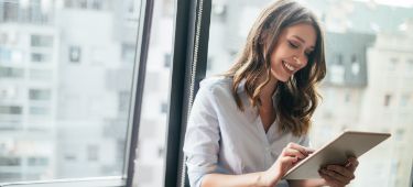 Junge Frau in Bürokleidung steht an einer Fensterfront und hält ein Tablet in der Hand.