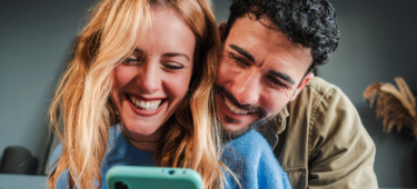 Paar bestehend aus Mann und Frau sitzen auf dem Sofa und blicken gemeinsam freudig auf ein Smartphone.