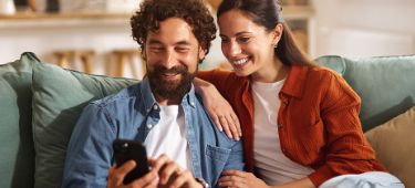 Ein Paar sitzt auf dem Sofa und schaut auf ein Handy. 