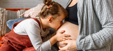 Große werdende Schwester im Schulkindalter küsst liebevoll Babybauch der Mutter.