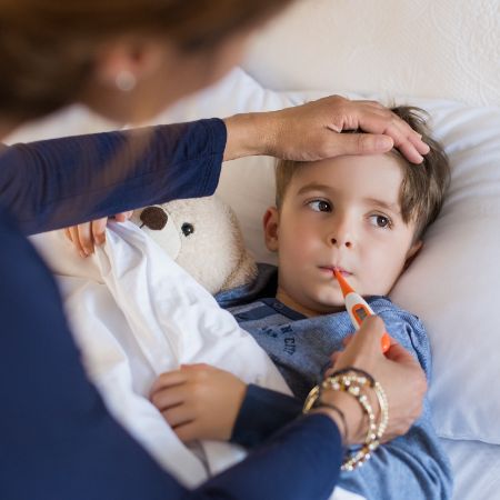 Ein kleiner Junge liegt krank im Bett und seine Mutter misst bei ihm Fieber.
