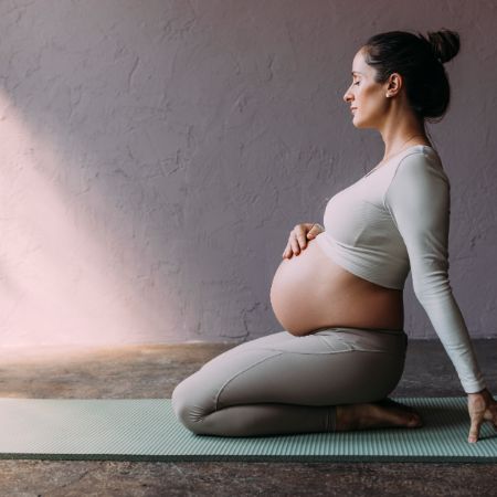 Eine schwangere Frau sitzt auf den Knien auf einer Gymnastikmatte und hält die Hand auf ihren Bauch.