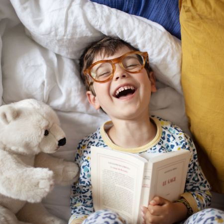 Lachender Junge mit Brille liegt im Bett und liest ein Buch.