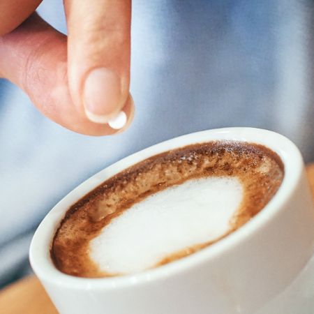 Zwei Finger werfen Suessstoff-Tablette in Kaffeetasse.