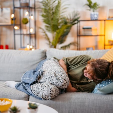Eine junge Frau liegt zusammengekrümmt vor Bauchschmerzen auf dem Sofa.