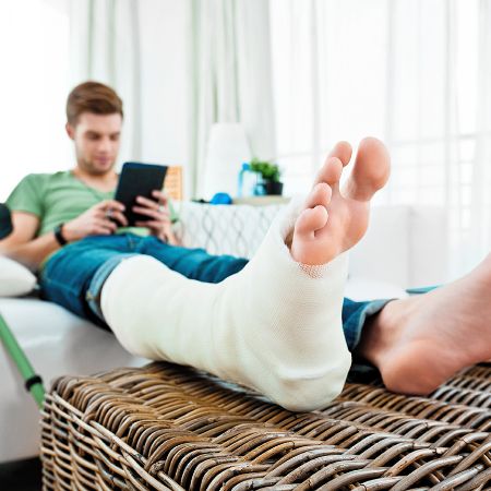 Mann mit Gipsbein und Krücken daneben liegt auf dem Sofa und liest ein e-Book.