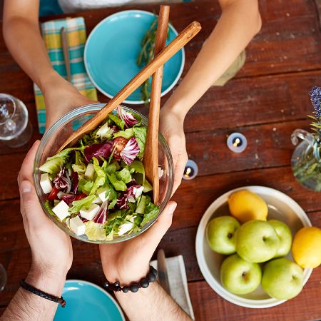 Zwei Hände halten eine Salatschüssel über dem Esstisch.